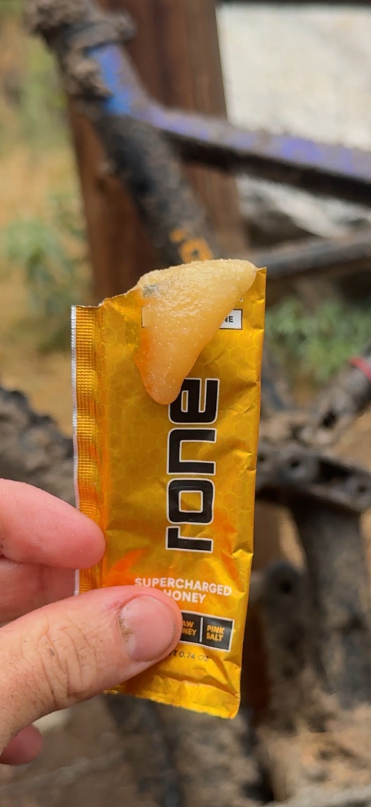 Hand holding an open RONE sachet of raw honey in front of a muddy mountain bike